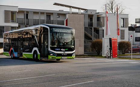 In einer ruhigen Stadtrandlage parkt ein nagelneuer Elektrobus an seiner Ladestation. Die urbane Umgebung mit minimalistischen Wohngebäuden und kahlen Winterbäumen vermittelt eine kühle, moderne Atmosphäre des öffentlichen Nahverkehrs im 21. Jahrhundert. Moderner elektrischer Stadtbus in grün-weißer Farbgebung an einer Ladestation vor zeitgenössischer Architektur.