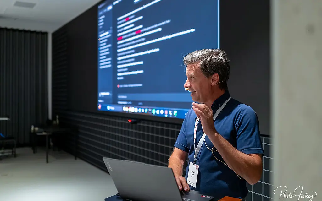 Prof. DDr. Roman Egger sits with a microphone and laptop in front of a large presentation screen. As digital content and programming code appear behind him, he explains current developments and practical applications of artificial intelligence.