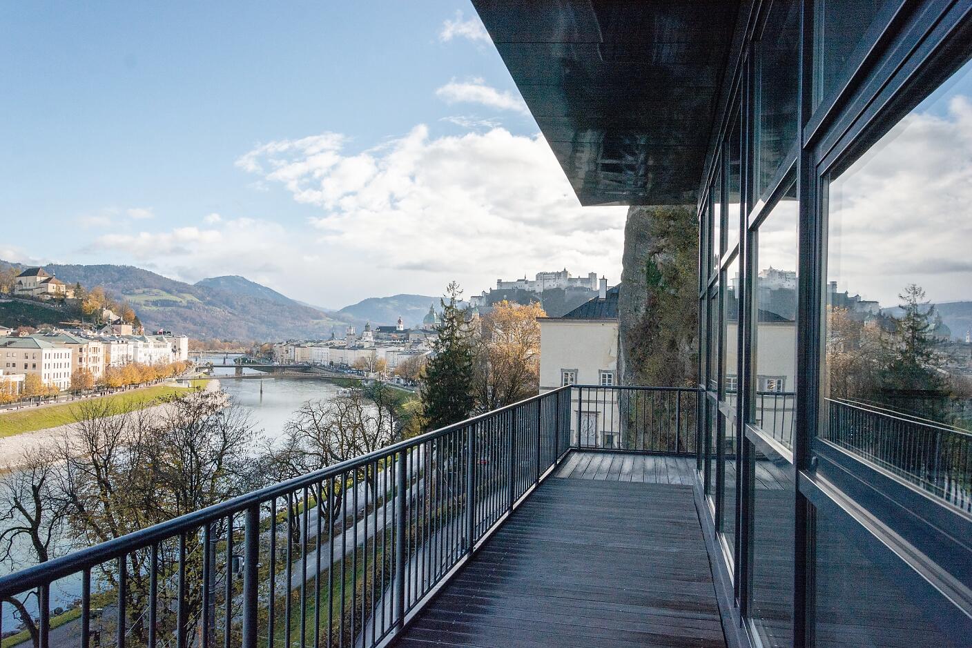 A breathtaking view unfolds from this glass balcony: the Salzach River winds through the autumnal cityscape, with the iconic Hohensalzburg Fortress rising majestically in the background. Gentle mountains and golden trees frame this picturesque scene, lending the moment a timeless beauty. Modern balcony overlooking the Salzach River and historic Salzburg's old town, framed by autumn trees and mountains.