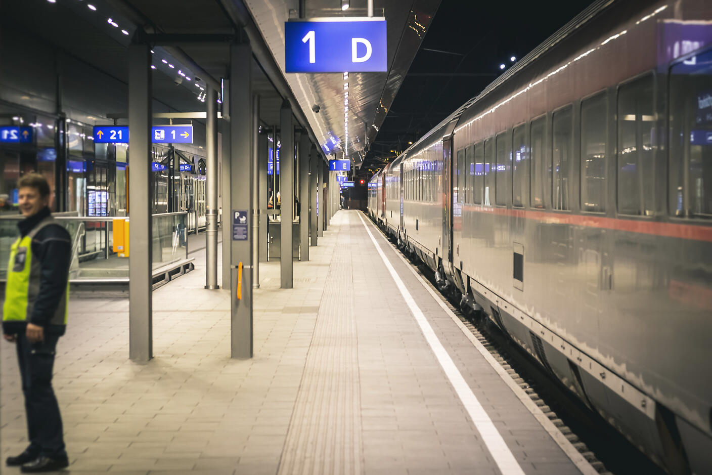 Öffentlicher Verkehr Salzburg Nächtlicher Bahnsteig mit blau beleuchteten Wegweisern, ein Zug steht auf Gleis 1D bereit. Ein Mitarbeiter in Warnweste steht am linken Bildrand.