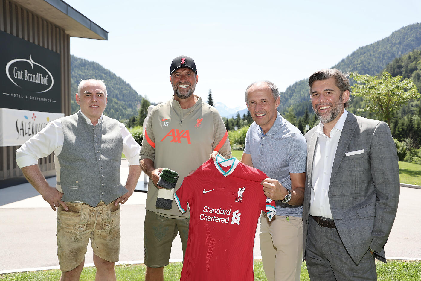 Against the picturesque backdrop of the SalzburgerLand, four gentlemen gather in a summery atmosphere. The mountainous landscape frames the scene, while a red and white football jersey serves as a connecting element between the men. The shot at Gut Brandlhof breathes a mix of professionalism and vacation mood. Four men pose in front of a mountainous landscape at Gut Brandlhof, with one person holding a red Liverpool jersey.