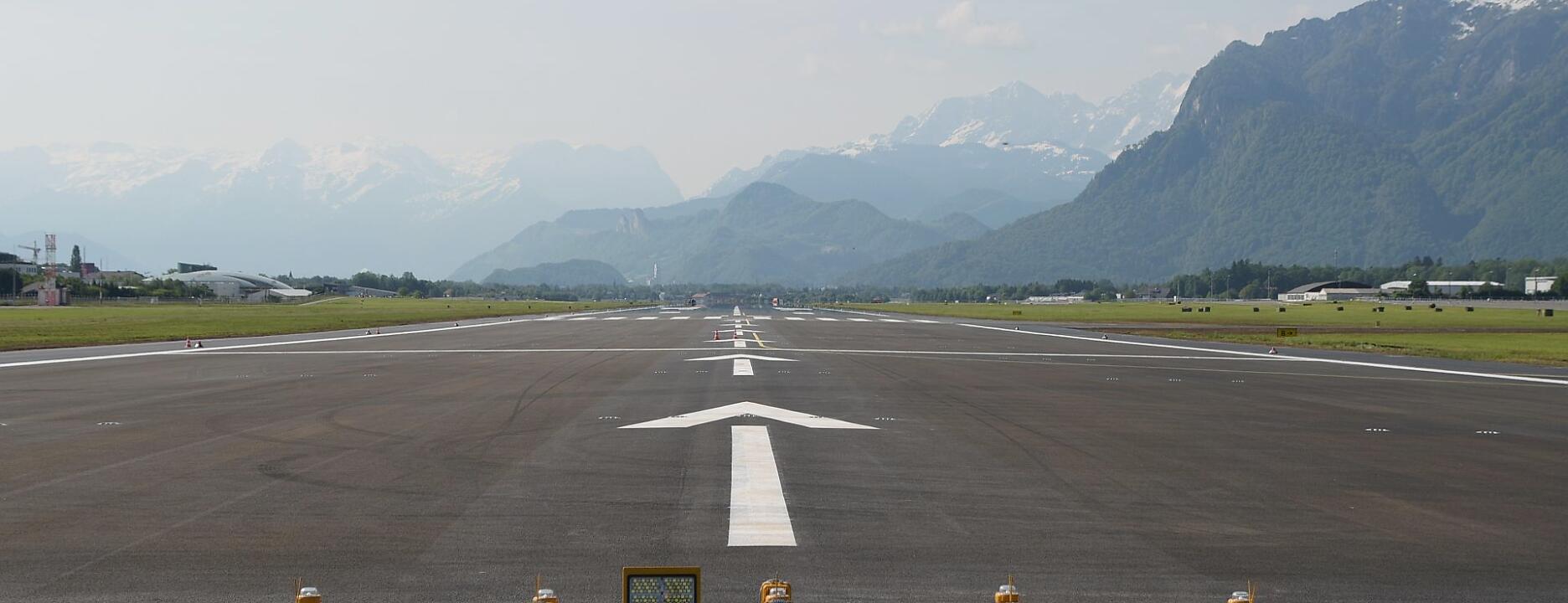 Die Startbahn des Salzburger Flughafens erstreckt sich geradeaus in die Ferne. Im Hintergrund erheben sich majestätisch die schneebedeckten Alpen, während das satte Grün der Umgebung in sanftem Kontrast zur markierten Asphaltfläche steht.