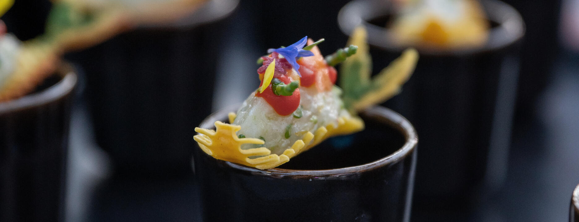 Artfully arranged finger food is served in elegant black porcelain cups. Vibrant edible flowers, fine vegetable strips, and a delicate chip create a visual and culinary masterpiece in miniature form. Colorful gourmet finger food with flowers and vegetables in black cups.