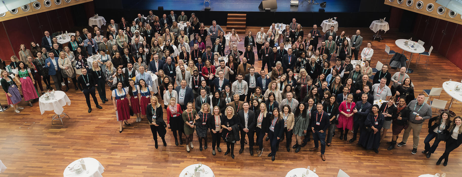 Große Gruppenaufnahme bei einer Konferenz mit vielen Teilnehmenden in einem modernen Konferenzsaal mit Holzboden und blauer Bühnendekoration.