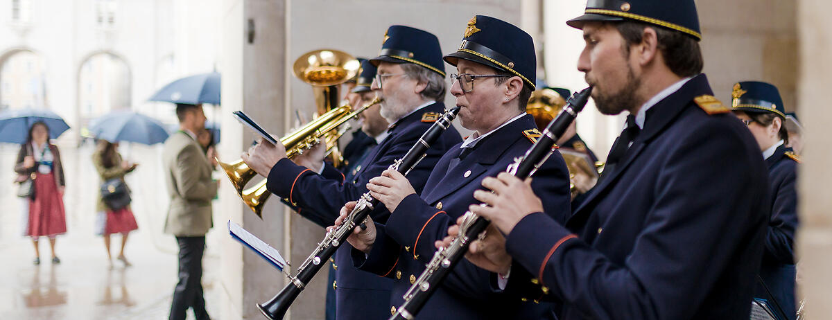 Blasorchester in blauen Uniformen spielt an einem regnerischen Tag vor historischer Architektur. Musiker mit Klarinetten und Trompeten stehen nebeneinander.