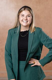 Ein Porträt voller Energie und Dynamik: Eine junge Frau strahlt in einem eleganten Business-Outfit aus dunkelgrünem Blazer und schwarzem Top. Mit einem sympathischen Lächeln und selbstbewusster Körperhaltung verkörpert sie moderne Professionalität und Lebensfreude. Lächelnde junge Frau in grünem Blazer und schwarzem Oberteil vor neutralem Hintergrund, selbstbewusst und professionell posiert.