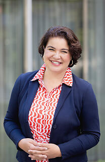 Vor einer modernen Glasfassade steht eine Frau mit kurzem Haar und einem offenen, herzlichen Lächeln. Ihr farbenfrohes Hemd und der dunkelblaue Blazer verleihen dem Porträt eine stilvolle, selbstbewusste Ausstrahlung in professionellem Umfeld.