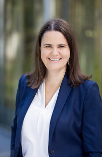 Vor einem modernen Glasgebäude steht eine lächelnde Frau in Businesskleidung. Ihr dunkelblauer Blazer und die weiße Bluse unterstreichen den professionellen Auftritt, während das natürliche Licht eine freundliche Atmosphäre schafft.