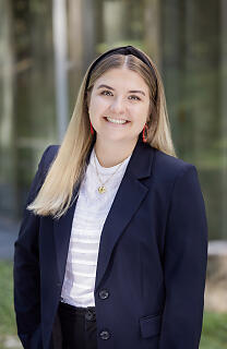 Junge Frau im Business-Outfit lächelt vor moderner Glasfassade selbstbewusst in die Kamera.