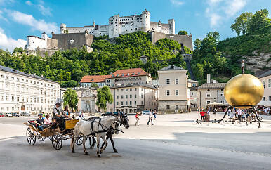 Historische Salzburger Altstadt mit der imposanten Festung Hohensalzburg auf einem bewaldeten Hügel, im Vordergrund eine Pferdekutsche und ein goldener Kunstball.