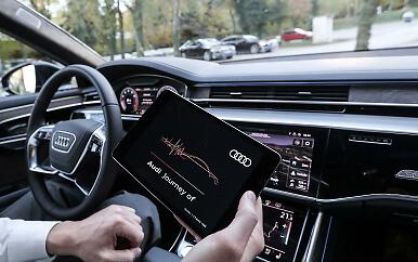 A modern vehicle interior reveals itself in technical perfection. The dashboard displays high-tech screens while a hand operates a tablet with an interactive Audi application. The atmosphere conveys high-tech elegance and driving refinement. Interior view of an Audi vehicle dashboard with a hand holding a tablet displaying 'Audi Journey of'.