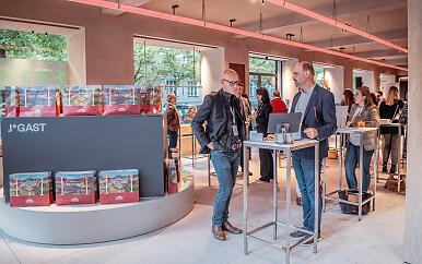 In a light-flooded room with reddish ceiling lighting, the Salzburg Convention Bureau booth presents itself professionally and invitingly. Elegantly dressed employees stand at high tables, surrounded by red and white welcome publications, conveying an atmosphere of hospitality and communicative openness. Modern trade fair booth of Salzburg Convention Bureau with welcome materials and two employees in a communicative setting.