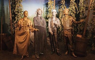 Eine surreale Szenerie entfaltet sich in einem goldenen Tableau: Vier stilisierte Figuren stehen in unterschiedlichen Posen, gehüllt in glänzende Gewänder, die sie wie lebendige Statuen erscheinen lassen. Die Hopfenranken und der verschwommene Hintergrund verleihen der Szene eine traumartige, museale Atmosphäre. Vier lebensgroße goldene Statuen in historischen Kostümen vor einer Kulisse aus Hopfenranken und Bäumen.