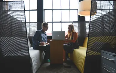 Zwei Personen in einer modernen Büro-Nische mit Sichtschutz, MacBook auf einem Holztisch und Stehlampe im Hintergrund.