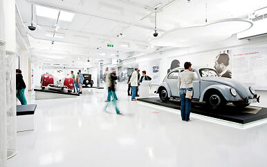 Modern white exhibition space with classic Volkswagen vehicles on pedestals, visitors exploring exhibits in minimalist architectural setting.