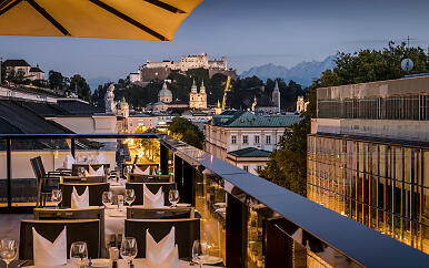On a stylishly set rooftop terrace, a breathtaking view opens up over Salzburg’s Old Town with the illuminated Hohensalzburg Fortress. The golden evening light lends the scene an elegant and romantic atmosphere. Rooftop terrace with views of Salzburg and Hohensalzburg Fortress at dusk.