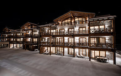 Inmitten einer winterlichen Schneelandschaft erstrahlt das Wildkogel Resort in sanftem Licht. Die harmonische Architektur verbindet rustikale Holzelemente mit modernen Steinakzenten und schafft eine einladende Atmosphäre für Winterurlauber. Mehrere Gebäudeebenen mit beleuchteten Balkonen und Fenstern versprechen Komfort und Gemütlichkeit in alpiner Bergkulisse. Nächtliche Aufnahme eines modernen Holz- und Steingebäudekomplexes im Winterdesign mit warmem Lichtschein aus den Fenstern und verschneiter Umgebung.
