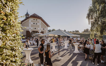 Sommerliche Outdoor-Veranstaltung mit weißen Stehtischen und Sonnenschirmen vor einem historischen Gebäude mit Turm, umgeben von Bäumen und Grünflächen.