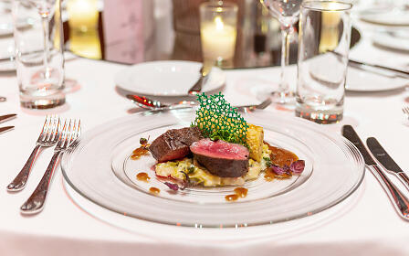 Elegante Gourmet-Hauptspeise auf weißem Teller: Rosa gebratenes Rinderfilet mit kunstvoller grüner Garnitur, umgeben von feiner Tischdekoration mit Kerzen und Weingläsern.