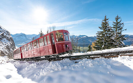 Red cogwheel train traversing a snowy mountain landscape with pine trees and mountain panorama under a bright blue winter sky.