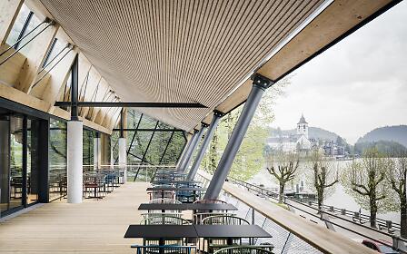 Modern wood and glass pavilion with panoramic view of a lake and a church, furnished with black tables and gray chairs.