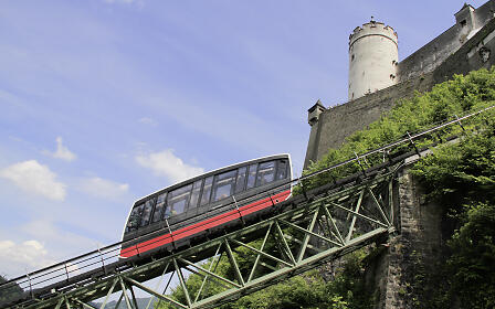 Funicular railway on a green steel bridge in front of a historic fortress wall with a white tower, against a blue summer sky.