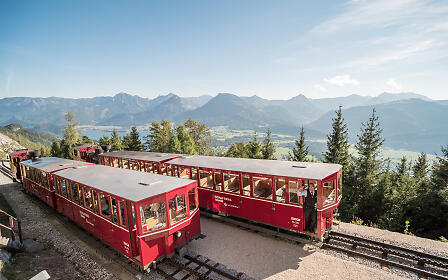 Rote Zahnradbahn auf einem Bergpass mit atemberaubender Aussicht auf eine alpine Gebirgslandschaft mit Tannen und weitläufigem Talblick.