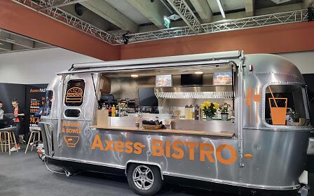 Silberner Foodtruck des Axess Bistros mit orangefarbener Beschriftung, ausgestattet für Burger und Bowls, in einer Messehalle.