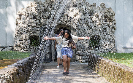 Two people on a narrow water path between stone walls with cascading water streams, featuring a woman with outstretched arms in the center.