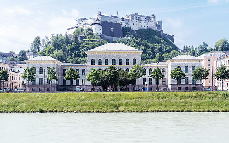 Die historische Universität Salzburg präsentiert sich in eleganter Architektur direkt am Ufer der Salzach. Im Hintergrund erhebt sich majestätisch die Festung Hohensalzburg über der Stadt und schafft gemeinsam mit der grünen Hügellandschaft ein beeindruckendes Panorama.