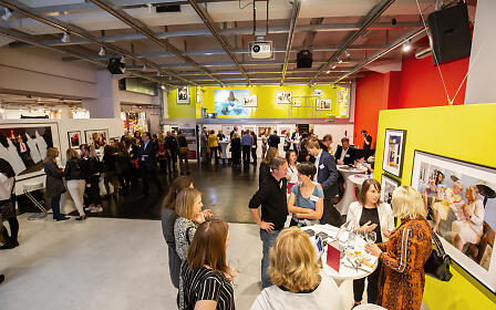 Kunstgalerie-Ausstellung mit zahlreichen Besuchern, die sich zwischen gerahmten Kunstwerken und gelb-roten Wänden bewegen und miteinander kommunizieren.