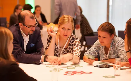 Three people sit at a white table, working intently with colorful elements during a meeting or workshop scenario.