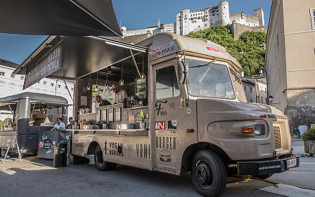 Ein cremefarbener Food Truck mit Vegan-Burger-Angebot steht vor historischer Architektur mit einer Festung im Hintergrund.