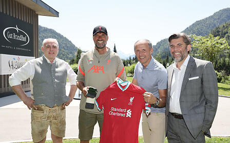 Four men pose in front of a mountainous landscape at Gut Brandlhof, with one person holding a red Liverpool jersey.