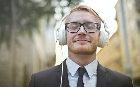 Ein Mann mit Brille und weißen Kopfhörern in einem Anzug, der entspannt mit geschlossenen Augen in urbaner Umgebung steht.