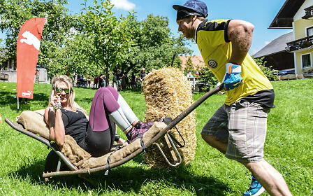 Sporty activity on a meadow: One person is being transported in a wheelbarrow by another person, surrounded by green landscape and buildings.
