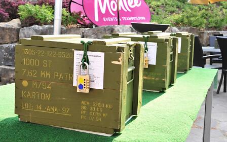 Olive green military ammunition boxes arranged on artificial green turf, set up for a team-building event.