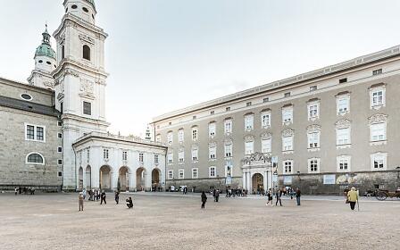 Historic square in Salzburg with baroque buildings, white church with green towers, and adjacent arcades, with scattered visitors moving about.