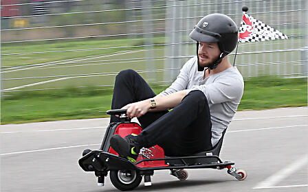 A person in a gray shirt and black pants rides a go-kart-like vehicle with a checkered flag on an asphalt surface with a fence and grass in the background.