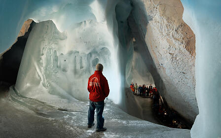 A person in a red jacket stands in a massive ice cave with impressive ice formations and a group of visitors in the background.