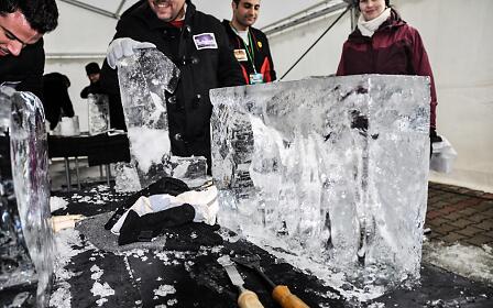 Inside a sheltered tent, participants carefully carve large ice blocks into impressive sculptures. Wearing gloves and using chisels with skill and precision, they shape fascinating structures from the transparent material as interested spectators watch the creative process unfold.