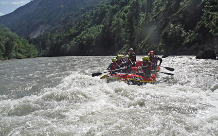 White water rafting on a turbulent river surrounded by dense forest-covered mountain slopes in a dramatic alpine landscape.