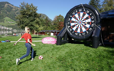 A person in a red shirt plays soccer dart on a green lawn with mountains and a lake in the background.