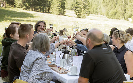 Outdoor wine tasting with a group of people at a set table against a green forest backdrop.