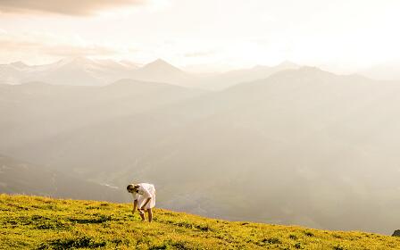 A person in a white dress bends down on a golden alpine meadow, with soft mountain silhouettes in warm sunlight in the background.