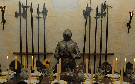 Mittelalterliche Ritterrüstung zwischen historischen Hellebarden, umgeben von gedeckter Tafel mit Kerzen und Sonnenblume in festlicher Atmosphäre.