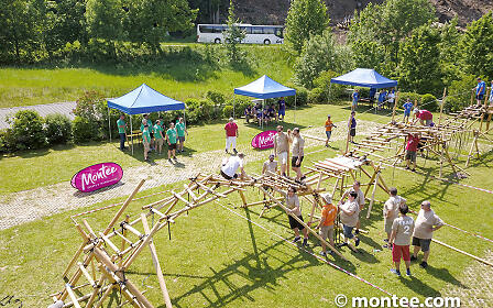 Team building event with wooden constructions on a green meadow. Blue pavilions and participants collaboratively working on a wooden framework project outdoors.