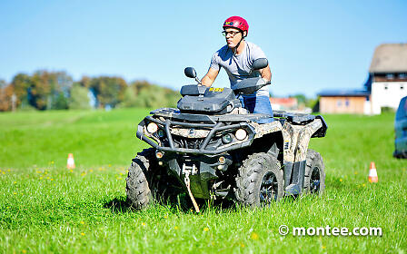 An ATV rider with a red helmet drives across a green meadow with rural buildings in the background.