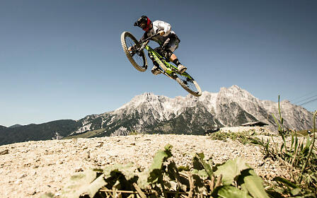 Ein Mountainbiker macht einen Sprung und fliegt gerade mit dem Bike in der Luft. Das Wetter ist sonnig und schön.