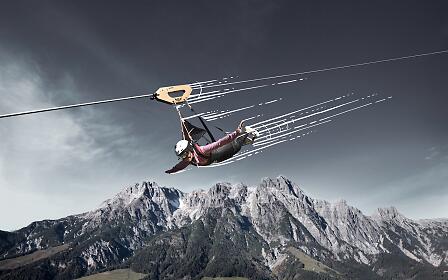 An adventurer speeds through the air on the Flying Fox XXL above the dramatic mountain landscape of Saalfelden Leogang. Secured to a steel cable, the rider glides high above green alpine meadows and rugged peaks, creating a thrilling experience with breathtaking views of the Alps.